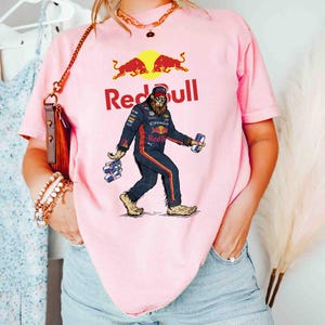 Pu&ograve; includere: T-shirt rosa chiaro con un personaggio simile a Bigfoot in tuta da corsa, con lattine di Red Bull. Il logo e il testo Red Bull sono sopra la grafica. La maglietta &egrave; di colore rosa chiaro.