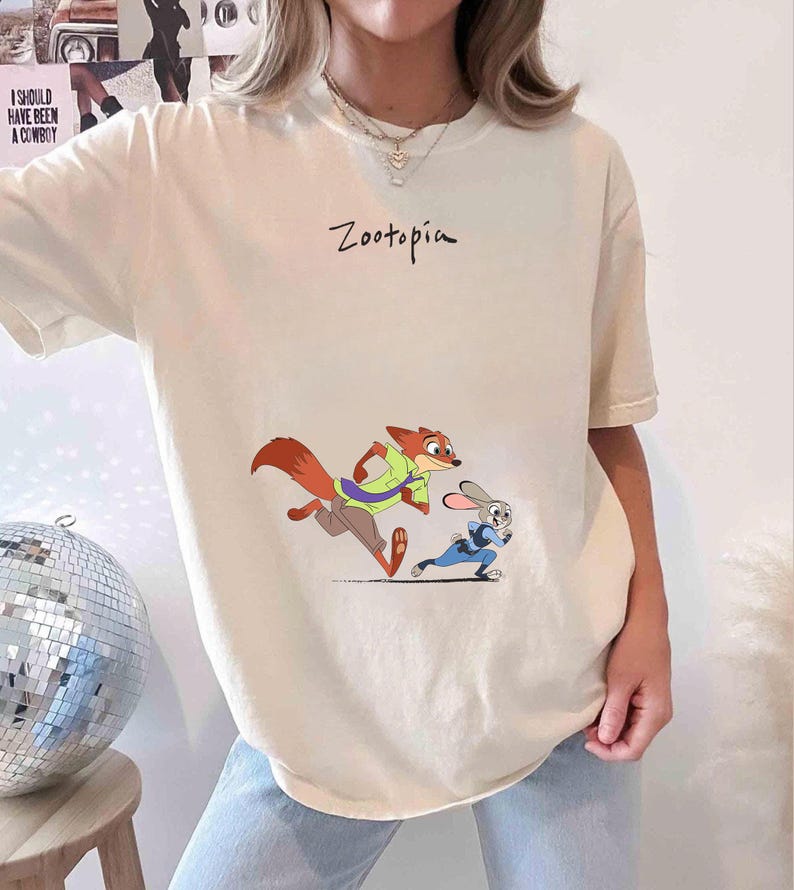 May include: Cream-colored t-shirt featuring the word "Zootopia" in black script. The shirt displays a cartoon image of a fox and a rabbit running. The fox is wearing a green shirt and the rabbit is wearing a blue uniform.