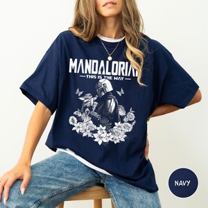 Może przedstawiać: Granatowa koszulka z napisem "MANDALORIAN" w białym kolorze nad ilustracją Mandalorianina i Grogu otoczonych białymi kwiatami i motylami. Tekst "THIS IS THE WAY" znajduje się pod słowem "MANDALORIAN".