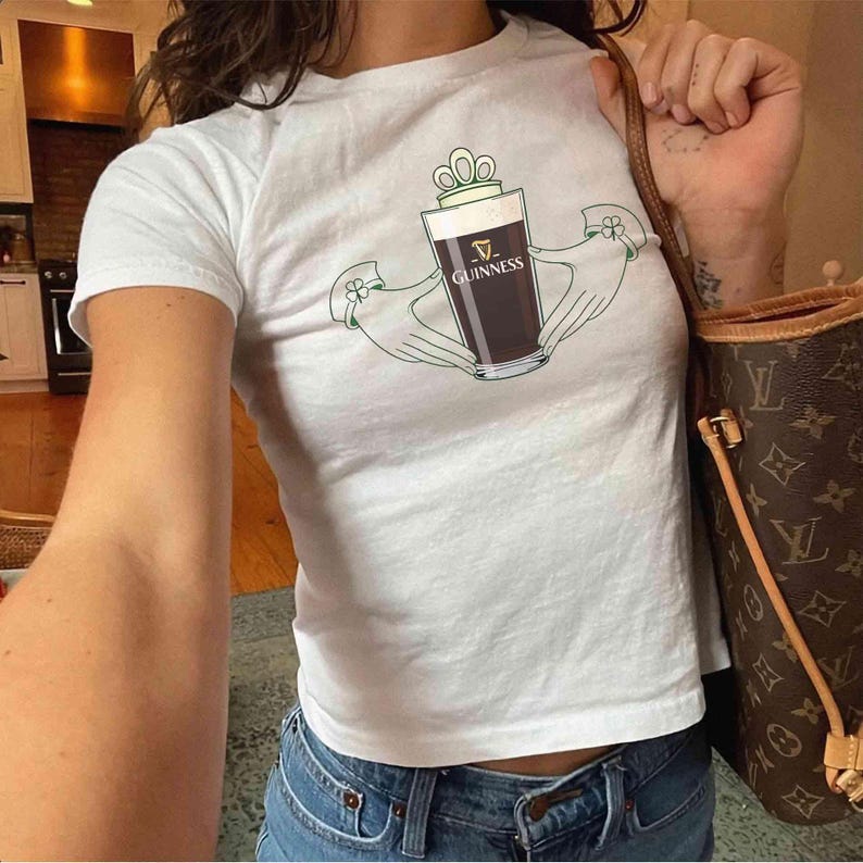 May include: White t-shirt featuring a graphic of a pint of Guinness stout held by two green hands, with the Guinness logo visible. The shirt is paired with blue jeans. A brown handbag with a monogram pattern is also visible.