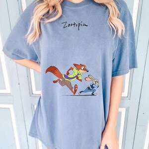 May include: A blue t-shirt featuring the word "Zootopia" in black script above a cartoon illustration of Nick Wilde and Judy Hopps running. Nick is a fox wearing a green shirt and Judy is a rabbit in a blue uniform.