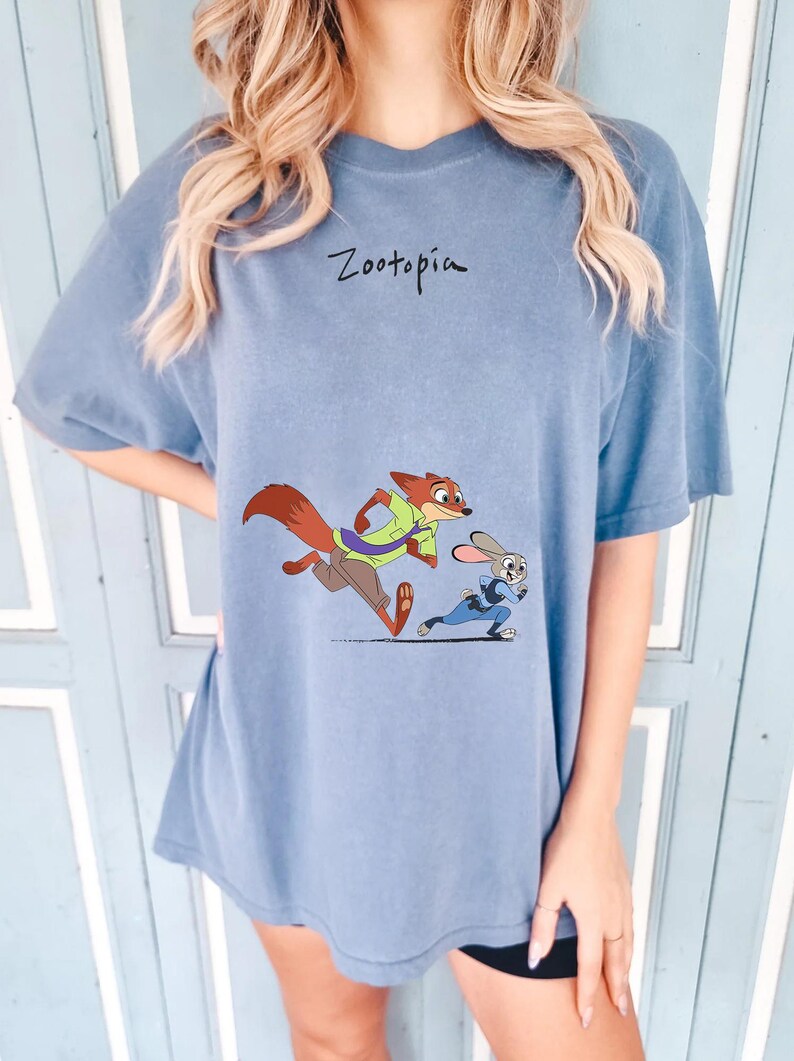 May include: A blue t-shirt featuring the word "Zootopia" in black script above a cartoon illustration of Nick Wilde and Judy Hopps running. Nick is a fox wearing a green shirt and Judy is a rabbit in a blue uniform.