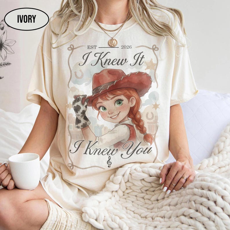 May include: An ivory short-sleeve t-shirt featuring a graphic of a cartoon cowgirl with red hair and a brown hat. The text reads "EST 2026", "I Knew It", and "I Knew You" with a musical note symbol. The design includes a decorative border.