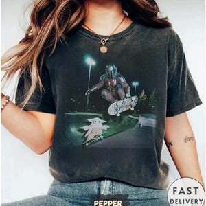 May include: Dark gray t-shirt featuring a graphic of The Mandalorian and Grogu skateboarding in a skate park. The Mandalorian is wearing his signature armor, and Grogu is riding a skateboard. The shirt has a vintage, worn look.