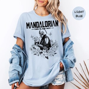 Może przedstawiać: Jasnoniebieska koszulka z napisem "MANDALORIAN" i frazą "THIS IS THE WAY". Grafika przedstawia czarno-białą ilustrację postaci Mandalorianina trzymającej małe stworzenie, otoczoną kwiatami i motylami.