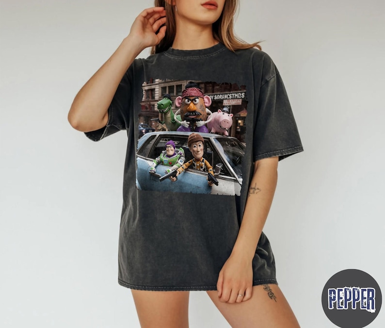 May include: A charcoal gray short-sleeve t-shirt featuring a graphic print of characters from Toy Story, including Buzz Lightyear, Woody, Mr. Potato Head, Rex, and Hamm, positioned in and around a blue car with a city background.