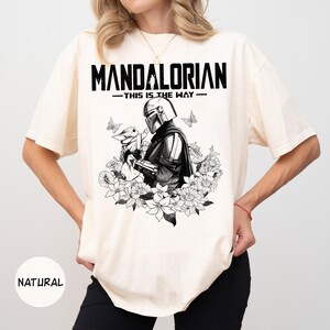 Może przedstawiać: Kremowa koszulka z czarno-białą grafiką Mandalorianina trzymającego Grogu, otoczonego kwiatami i motylami. Tekst "MANDALORIAN - THIS IS THE WAY" jest wydrukowany u g&oacute;ry pogrubionymi, czarnymi literami.