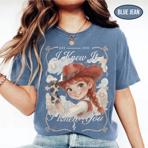 May include: A blue jean colored t-shirt featuring a graphic of a cartoon cowgirl with red hair in a braid, wearing a brown hat and cow-print glove. The text reads EST 2026, I Knew It, and I Knew You, framed by a rope design with stars.