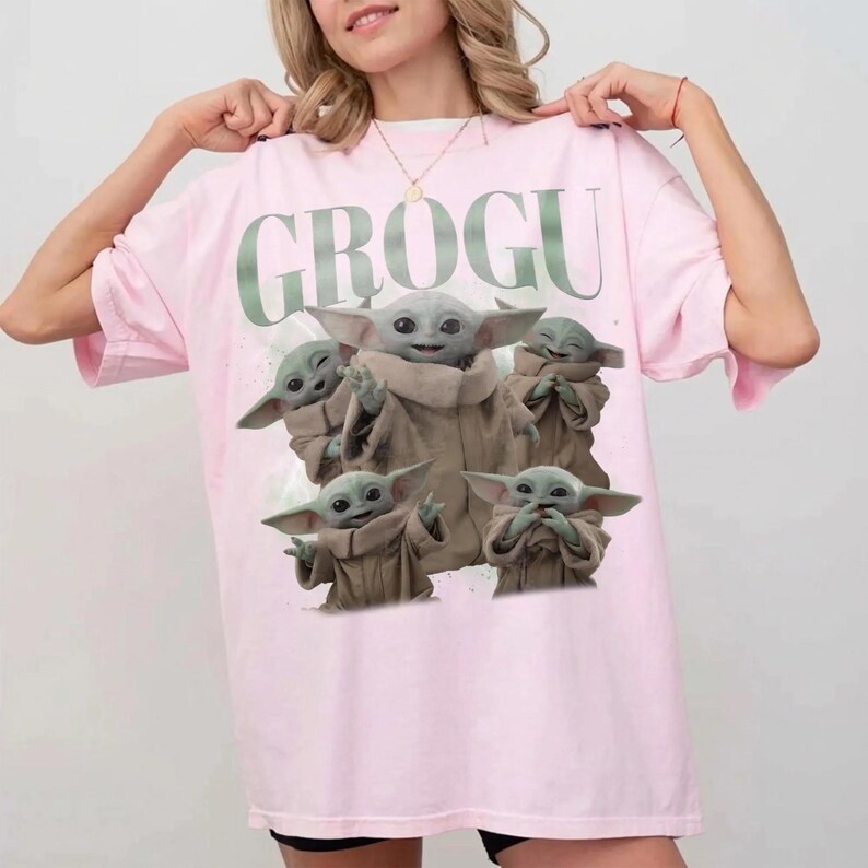 May include: A light pink t-shirt featuring the word "GROGU" in green above a graphic of Grogu from The Mandalorian. The graphic shows Grogu in a brown cloak with large ears, surrounded by other images of Grogu.