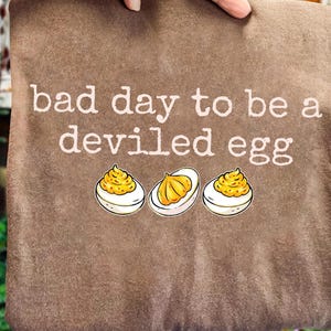 Può includere: Cuscino marrone con la scritta bianca "bad day to be a deviled egg". Sotto, tre illustrazioni di uova alla diavola con ripieno giallo e albumi bianchi. Il cuscino è tenuto da una mano.