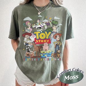 以下が含まれることがあります： モスグリーンのTシャツ。アニメ映画「トイ・ストーリー」のキャラクターのカラフルなグラフィックが特徴です。デザインにはウッディ、バズ・ライトイヤー、その他のキャラクターが含まれており、「TOY STORY」と「YOU'VE GOT A FRIEND IN ME」の文字があります。