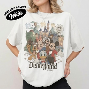May include: A white t-shirt featuring a vintage-style graphic of Mickey Mouse, Minnie Mouse, Donald Duck, and Pluto in front of a castle and Ferris wheel. The text "Disneyland" and "Est. 1955" is printed below the characters in a dark serif font.