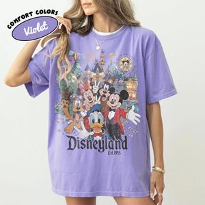 May include: A violet short-sleeve t-shirt featuring a graphic print of Mickey Mouse, Minnie Mouse, Donald Duck, Goofy, and Pluto in front of a castle. The text "Disneyland Est. 1955" is printed below the characters. A "Comfort Colors Violet" label is visible.