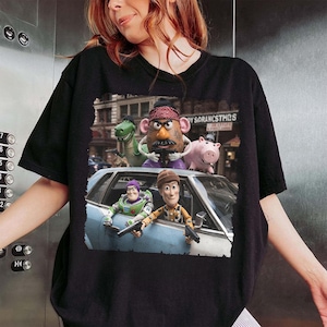 May include: A black t-shirt featuring a graphic print of characters from the Toy Story franchise, including Buzz Lightyear, Woody, Mr. Potato Head, Rex, and Hamm, depicted in a gritty, cinematic scene with a light blue car and a city background.