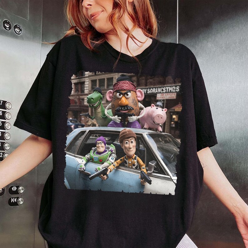 May include: A black t-shirt featuring a graphic print of characters from the Toy Story franchise, including Buzz Lightyear, Woody, Mr. Potato Head, Rex, and Hamm, depicted in a gritty, cinematic scene with a light blue car and a city background.