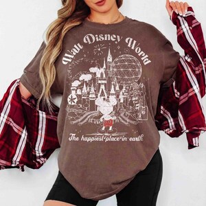 May include: Brown t-shirt with a white graphic of the Walt Disney World logo, including Cinderella's castle, Spaceship Earth, and Mickey Mouse. The text reads "Walt Disney World" and "The happiest place on earth."