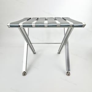 Pode incluir: Um suporte dobrável para bagagem com uma estrutura de metal prateado e quatro tiras de tecido cinzento dispostas horizontalmente no topo. A estrutura é apoiada por pernas em forma de X com pés arredondados para uma base estável.