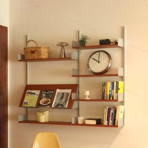 May include: A wooden wall-mounted shelving unit with several shelves. The shelves display books, magazines, a clock, a basket, and decorative lamps. The unit is attached to the wall with silver metal supports. The clock has a diameter of approximately 30 cm.
