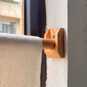 Może przedstawiać: Drewniany uchwyt na karnisz zamontowany na białej ścianie. Uchwyt wykonany jest z jasnego drewna i posiada cylindryczny drążek oraz kwadratowy wspornik. Kremowa zasłona wisi na drążku, częściowo zasłaniając okno.