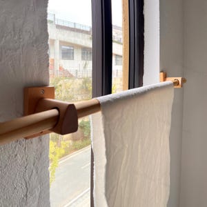Op de afbeelding: Een houten handdoekenrek met een lichte afwerking is aan een witte muur bevestigd. Een witte handdoek hangt aan het rek. Het rek bevindt zich voor een raam met uitzicht naar buiten.
