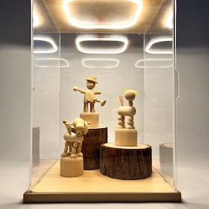 May include: A display case featuring three wooden figurines. The figurines are displayed on wooden platforms inside a clear acrylic box with a wooden base and top. The case is illuminated with a warm, soft light.