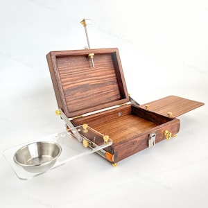 Könnte beinhalten: Eine Künstler-Malbox aus dunklem Holz mit einem Scharnierdeckel und einer kleinen, befestigten Metallschale. Die Box hat Messingbeschläge und eine transparente Acrylpalette. Die Box ist geöffnet und zeigt einen leeren Innenraum.