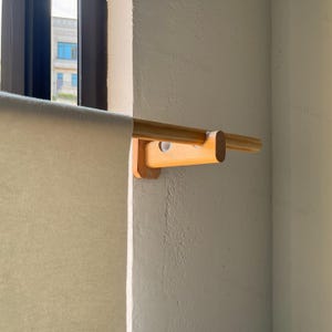 Puede incluir: Un soporte de madera de tono claro para barra de cortina montado en la pared, con una base en forma de U que sostiene una barra cilíndrica de madera a juego. Una pieza de tela lisa de color blanco roto cuelga de la barra, ocultando parcialmente la ventana.