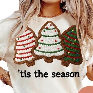 Puede incluir: Sudadera color crema con tres diseños de árboles de Navidad de ganchillo con bordes marrones. Los árboles están decorados con detalles rojos, verdes y blancos. La frase "'tis the season" está impresa en negro en la parte inferior de la camisa.