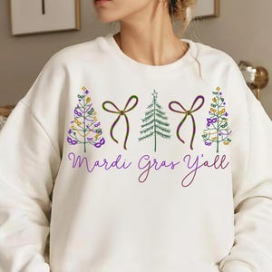 Può includere: Felpa color crema con tema Mardi Gras. Il design include alberi stilizzati, fiocchi e maschere in verde, viola e oro. Il testo "Mardi Gras Y'all" è scritto in corsivo.