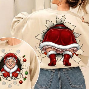 May include: Beige sweatshirt with a graphic of Santa Claus's backside bursting through a torn wall. The Santa graphic features a red suit with white fur trim and red boots. A smaller graphic of Mrs. Claus is on the front.