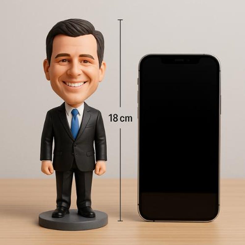 May include: A bobblehead figurine of a smiling man in a black suit and blue tie, standing 7 inches tall. The figurine is next to a black smartphone. The bobblehead is on a gray circular base.
