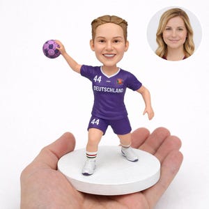 Könnte beinhalten: Eine Figur einer Frau in einem lila Handball-Trikot mit dem Wort "Deutschland" auf der Vorderseite. Die Figur hält einen lila und rosa Handball. Die Nummer "44" steht auf Trikot und Shorts. Die Figur steht auf einem weißen Sockel.