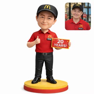 Puede incluir: Figura de un empleado de McDonald's con polo rojo, pantalón negro y gorra negra con el logo de McDonald's. La figura sostiene un letrero que dice "20 AÑOS!" y hace una señal de aprobación. La base es amarilla y roja.