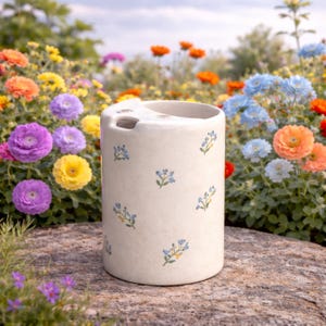 Puede incluir: Un portacepillos cilíndrico de cerámica blanquecina con la parte superior curvada y pequeños diseños florales azules. El soporte está sobre una superficie de piedra, rodeado de flores coloridas en un jardín.