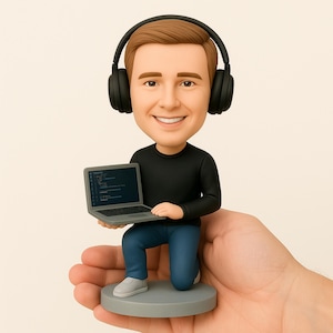 May include: A figurine of a smiling person wearing headphones, black long-sleeve shirt, and blue jeans, kneeling on a gray base. The figurine holds a gray laptop displaying code. The figurine is being held in a hand.