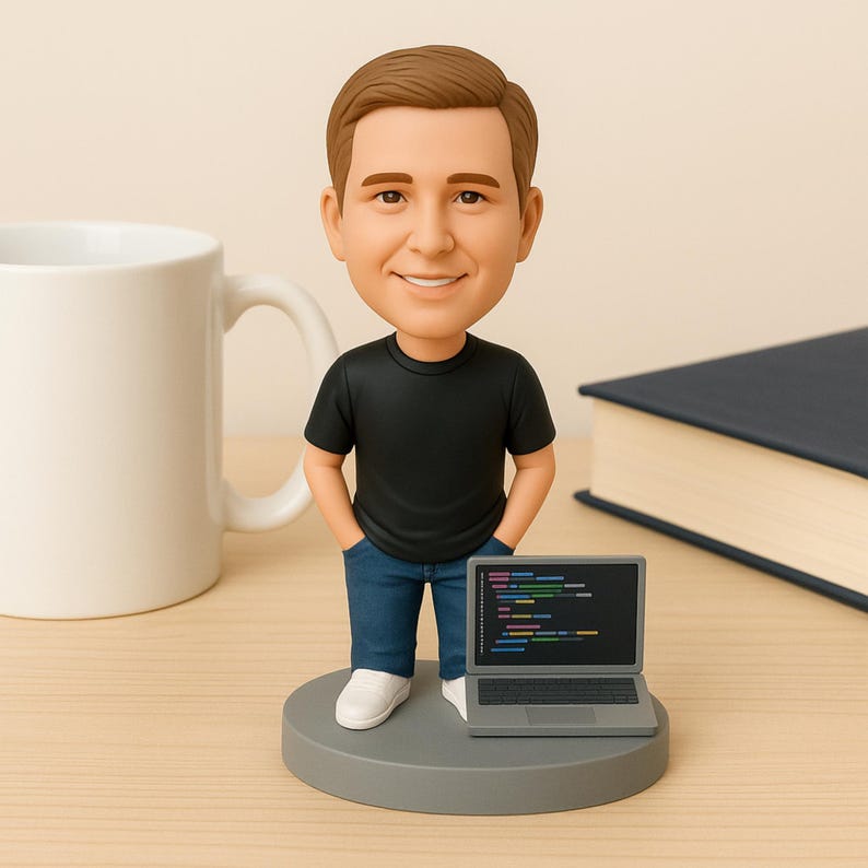 May include: A figurine of a man in a black t-shirt and blue jeans, with hands in his pockets, standing on a gray base. A small laptop with a colorful code display sits in front of him. A white mug and a book are in the background.