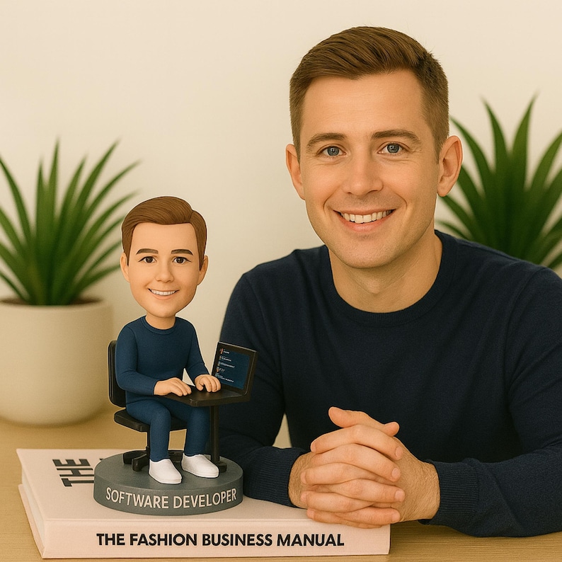 May include: A bobblehead figurine of a software developer sitting at a desk with a laptop. The figurine is on a base that says "SOFTWARE DEVELOPER". The figurine is sitting on a book titled "THE FASHION BUSINESS MANUAL". A man in a navy blue shirt smiles in the background.
