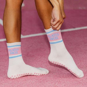 May include: White Pilates socks with pink and blue stripes at the top, featuring the text "PILATES ANGEL". The soles have pink heart-shaped grips. The socks are worn on a pink surface.