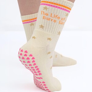 May include: Cream-colored socks with the text "The Life of a Barre Girl" in orange. The socks have pink and orange stripes at the top, gold star accents, and pink heart-shaped grips on the soles. The socks are designed for barre workouts.