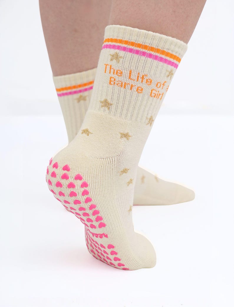 May include: Cream-colored socks with the text "The Life of a Barre Girl" in orange. The socks have pink and orange stripes at the top, gold star accents, and pink heart-shaped grips on the soles. The socks are designed for barre workouts.