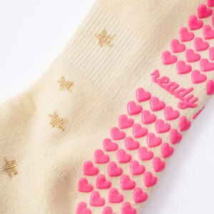 May include: Cream-colored sock with gold star patterns and pink heart-shaped grips. The word "ready" is written in pink on the side of the sock. The sock is made of a soft, ribbed material.