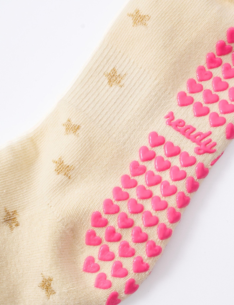May include: Cream-colored sock with gold star patterns and pink heart-shaped grips. The word "ready" is written in pink on the side of the sock. The sock is made of a soft, ribbed material.