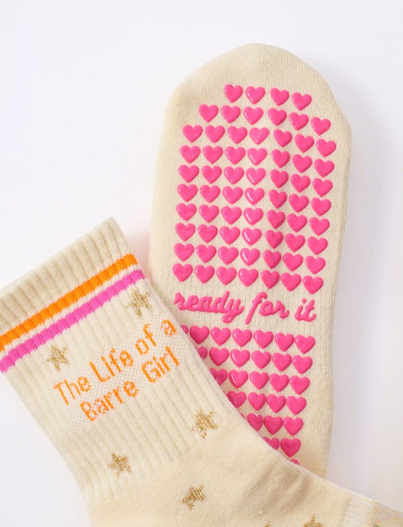 May include: Cream-colored socks with orange and pink stripes and the text "The Life of a Barre Girl." The soles feature pink heart-shaped grips and the words "ready for it." Gold star patterns are also visible.