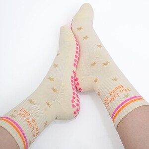 May include: A pair of cream-colored socks with gold star patterns and the text "The Life of a Barre Girl" in orange. The socks have pink hearts on the soles and orange and pink stripes at the top.