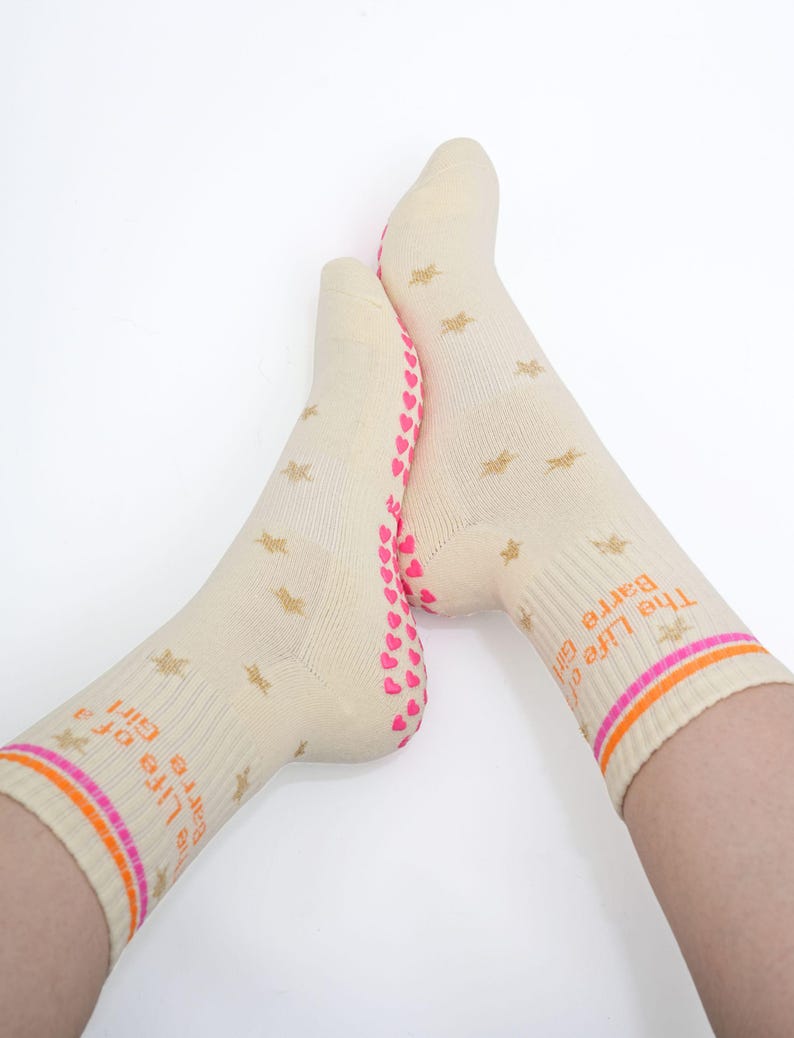 May include: A pair of cream-colored socks with gold star patterns and the text "The Life of a Barre Girl" in orange. The socks have pink hearts on the soles and orange and pink stripes at the top.
