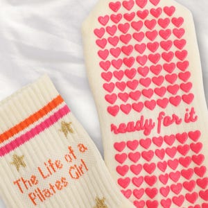 May include: Cream-colored Pilates socks with pink heart grips on the soles. The socks have orange and pink stripes and gold star accents. Text on the side reads "The Life of a Pilates Girl" and "ready for it".