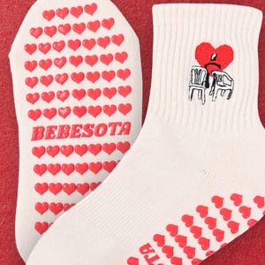 Könnte beinhalten: Weiße Socken mit roten herzförmigen Grips und dem Text "BEBESOTA" auf der Sohle. Eine Socke hat ein gesticktes Design eines roten Herzens mit einem traurigen Gesicht auf einem Stuhl. Die andere Socke hat den Text "SOTA".