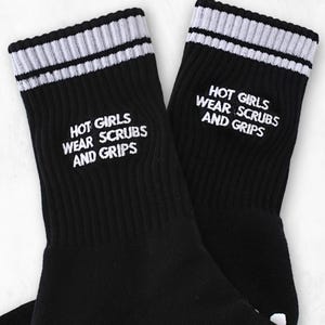 Könnte beinhalten: Schwarze Sportsocken mit weißem Text "HOT GIRLS WEAR SCRUBS AND GRIPS". Die Socken haben ein weiß-graues Streifenband am oberen Rand und weiße Griffe an den Sohlen. Die Socken sind gerippt.