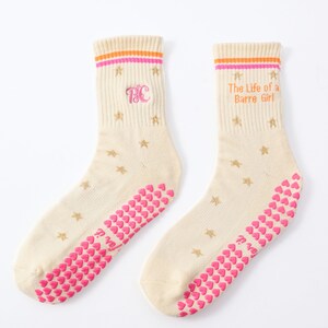 May include: A pair of cream-colored socks with pink and orange stripes at the top. The socks feature gold star patterns, pink heart-shaped grips on the soles, and the text "The Life of a Barre Girl". One sock has the letters "BC".