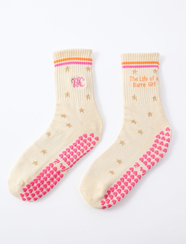 May include: A pair of cream-colored socks with pink and orange stripes at the top. The socks feature gold star patterns, pink heart-shaped grips on the soles, and the text "The Life of a Barre Girl". One sock has the letters "BC".